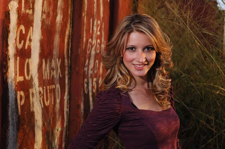 A young woman with curly blonde hair smiling near a rustic, weathered fence outdoors.