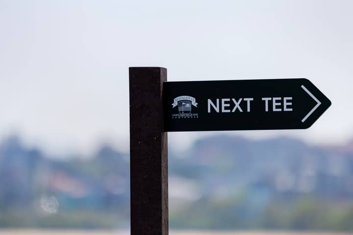 A directional sign on a golf course pointing to the next tee against a blurred background.