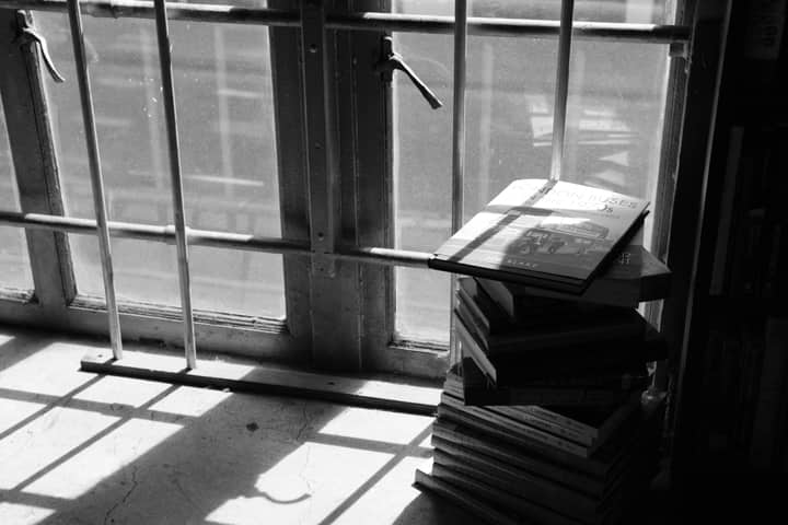 A stack of books sits by a barred window with sunlight casting shadows.