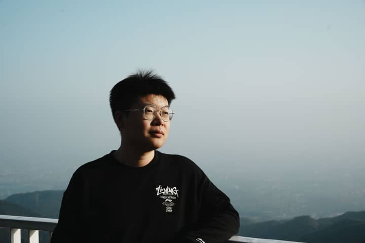 A man with glasses stands on a balcony, gazing at the mountain view under a clear blue sky.