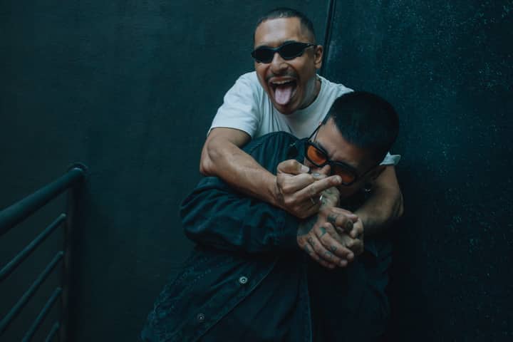 Two friends joking around indoors, wearing sunglasses and displaying playful gestures.