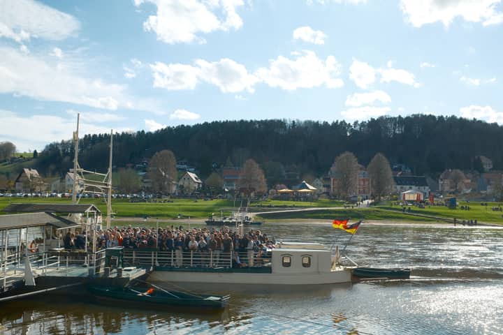 A ferry crosses a scenic river in Germany under a bright blue sky, filled with passengers.