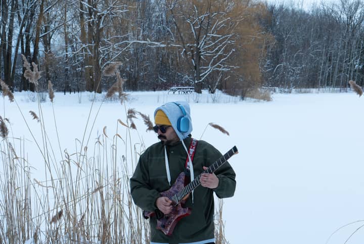 Man playing guitar in snowy park, Montreal, wearing headphones and winter clothes.