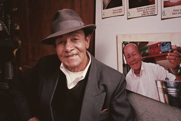 Elderly man wearing a hat smiles in front of a wall featuring a personal photograph in Quito, Ecuador.