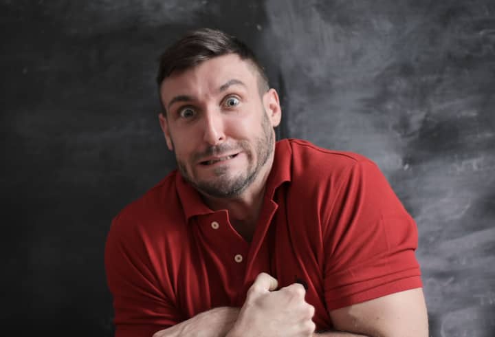 Man in red shirt making a humorous, exaggerated facial expression against a dark background.
