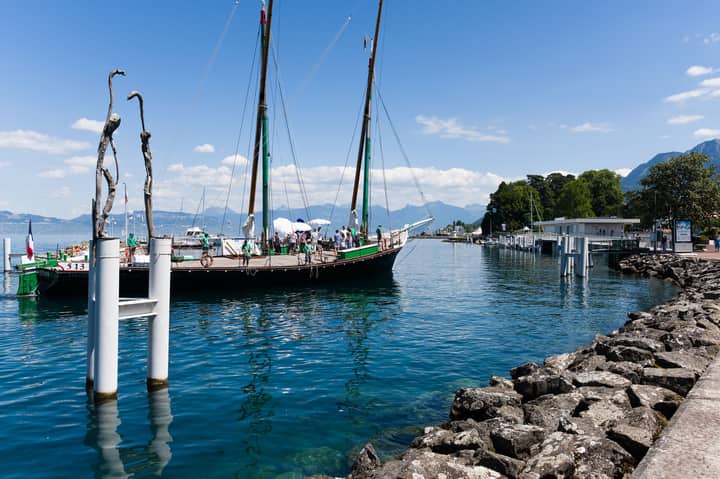 A traditional sailboat rests in the vibrant harbor of Evian, France, with the Alps in the background.