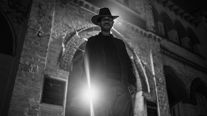 A man wearing a hat stands beneath a historic arch, backlit dramatically at night.