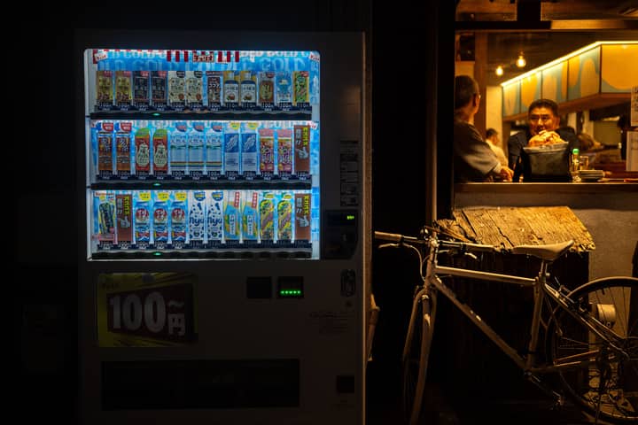 Vending machine glowing at night beside a cozy café with two men inside.