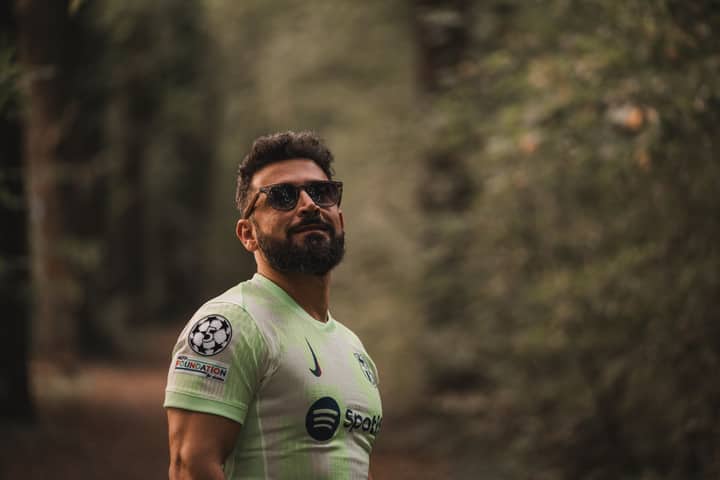 A bearded man in a soccer jersey stands confidently in a serene forest setting.