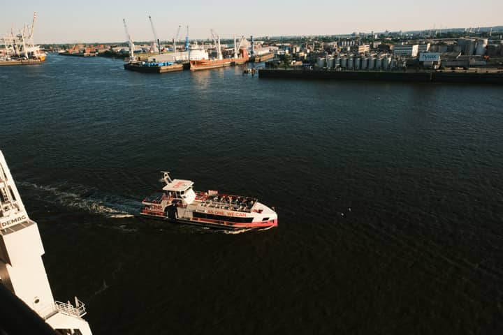 Aerial shot of a passenger boat cruising through an industrial harbor with cranes and ships under a clear sky.