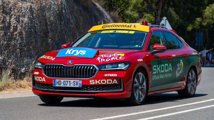 Red ŠKODA support car at Tour de France with branded decals on scenic route.