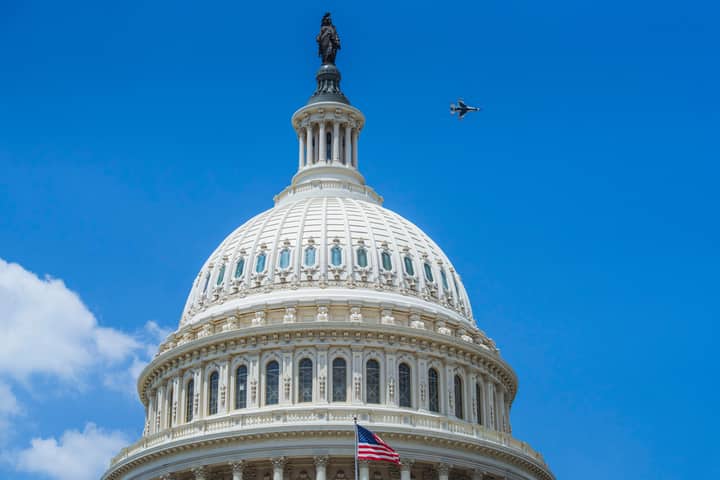 The US Capitol dome in Washington DC with a jet flying above under clear blue skies.