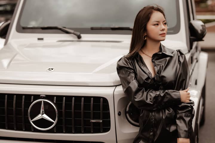 Fashionable woman in leather jacket leans on Mercedes G-Class, exuding urban elegance.