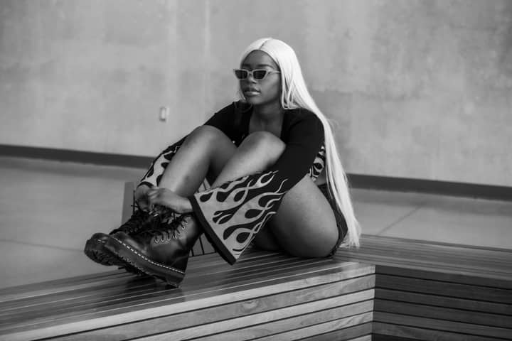 Black and white of young African American female with long white hair in trendy outfit and sunglasses relaxing on wooden bench