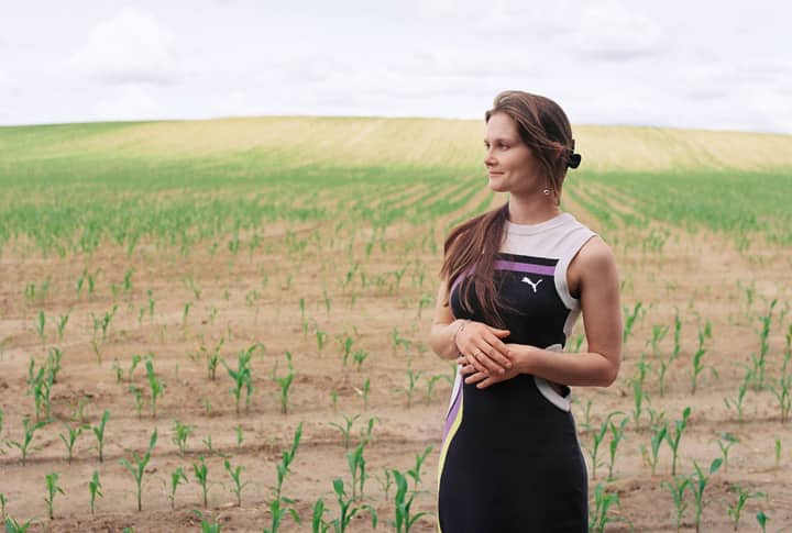 A young woman stands thoughtfully in a vast field under a bright summer sky.