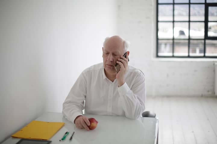 Serious male manager in formal clothes talking on phone while sitting at table with apple and documents in spacious room