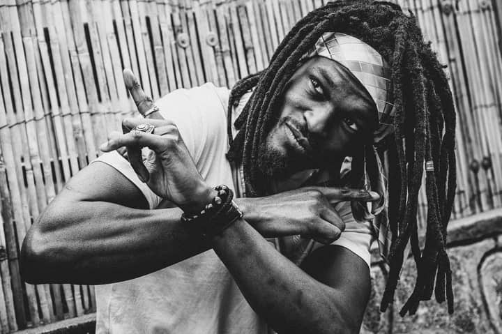 Black and white portrait of an adult man with dreadlocks, posing outdoors.
