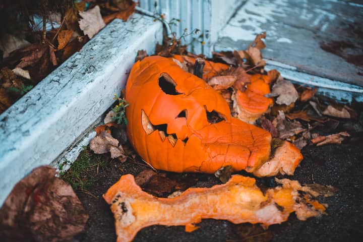 A smashed jack-o'-lantern surrounded by dry autumn leaves on a street corner.