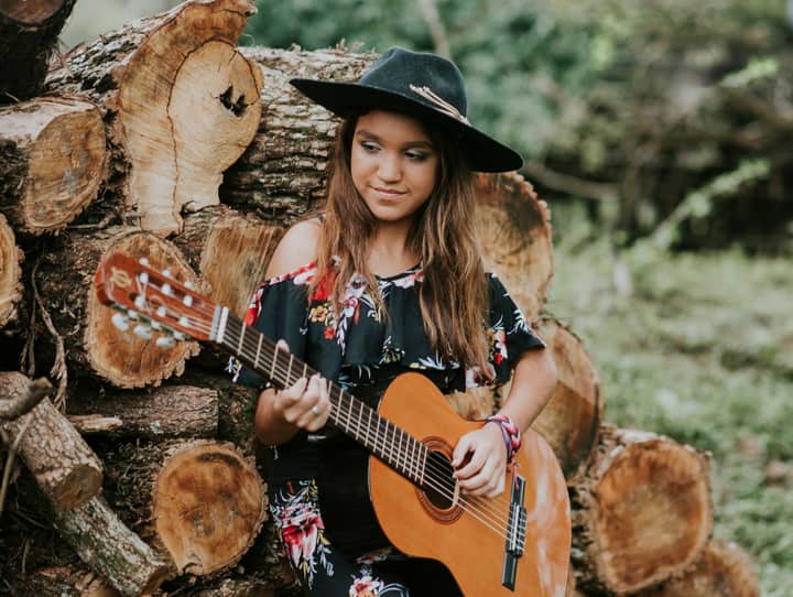 A woman in a floral dress and fedora serenely plays guitar by stacked logs.