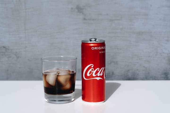 Chilled Coca-Cola can and drink with ice cubes in glass on minimalistic surface.