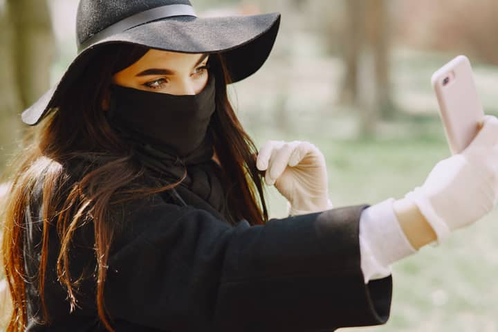 Crop female in stylish outerwear and medical mask and gloves taking selfie on smartphone in park during walk on city street