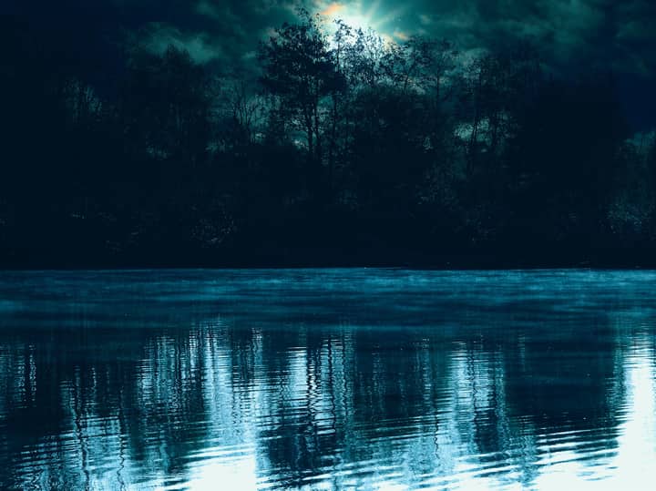 A dramatic dusk scene with silhouetted trees reflected in a tranquil lake under a cloudy sky.