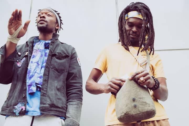 From below of African American hipster male with raised arm near partner with accessories and fabric bag standing near wall