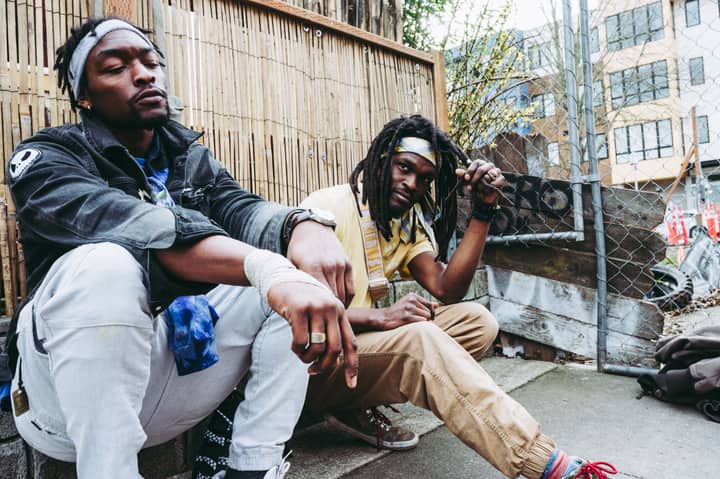 Serious African American men with dreadlocks wearing casual clothes sitting together on hunkers