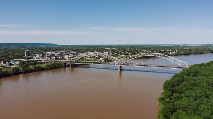 From above of modern metal bridge above brown river near trees and buildings under cloudy sky