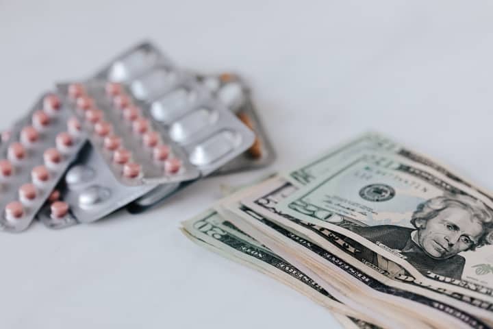 Heap of similar American dollars in cash placed near blisters of expensive drugs on white table