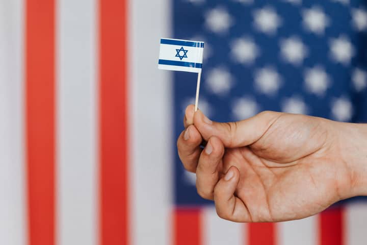 Closeup of crop unrecognizable person holding small flag of Israel before huge flag of United States of America on background