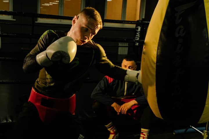 Focused boxer delivers a punch to a heavy bag under the watchful eye of a trainer.