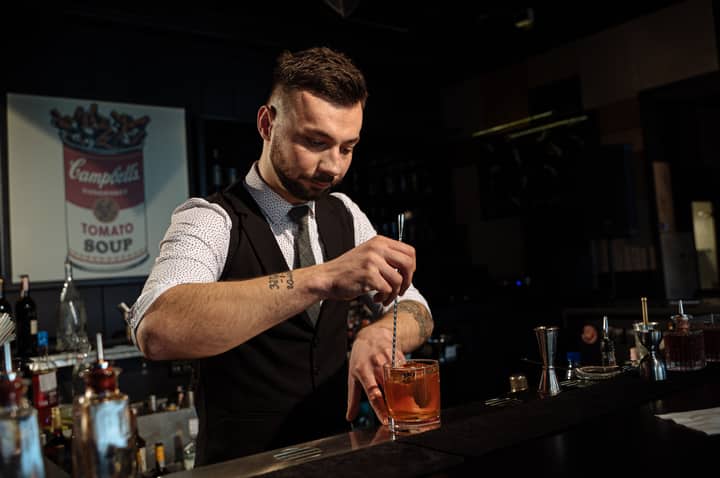 Bartender skillfully mixing a cocktail at a stylish bar with a Campbell's Soup poster in the background.