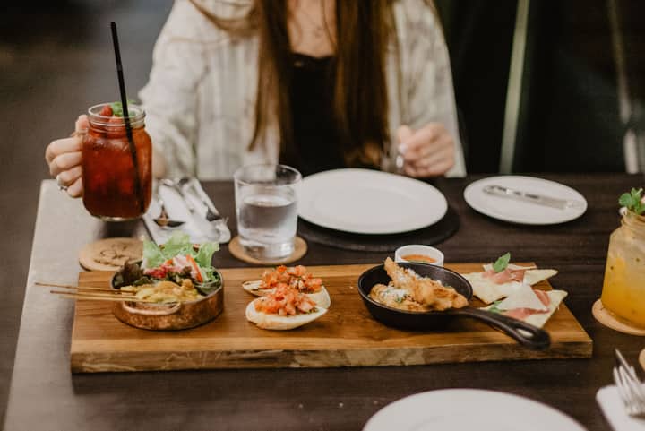 Crop unrecognizable female in casual clothes having dinner in Asian restaurant while sitting at table with berry mocktail and grilled meat on sticks served with tomato sandwiches and spring rolls