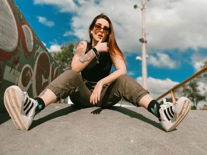 Low angle of confident young tattooed female in casual clothes and sunglasses touching lips and looking at camera while sitting on ground amidst walls with graffiti