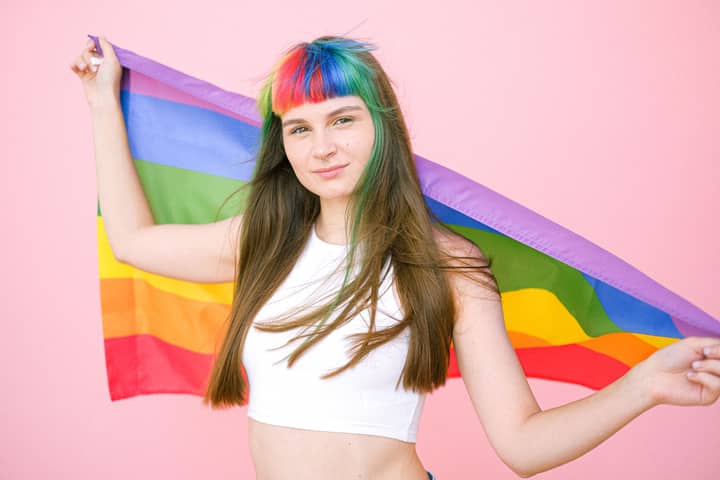 A woman with rainbow bangs holding a pride flag against a pink background, symbolizing LGBTQ pride.