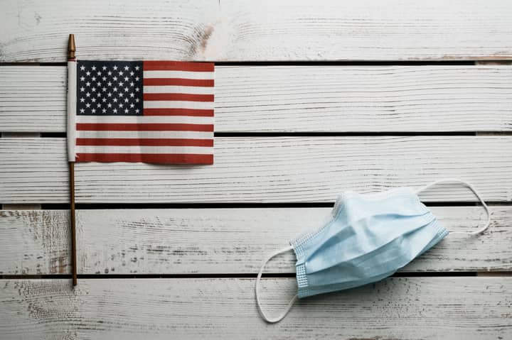 Top view of small Flag of United States and light blue fabric disposable face mask on white wooden wall
