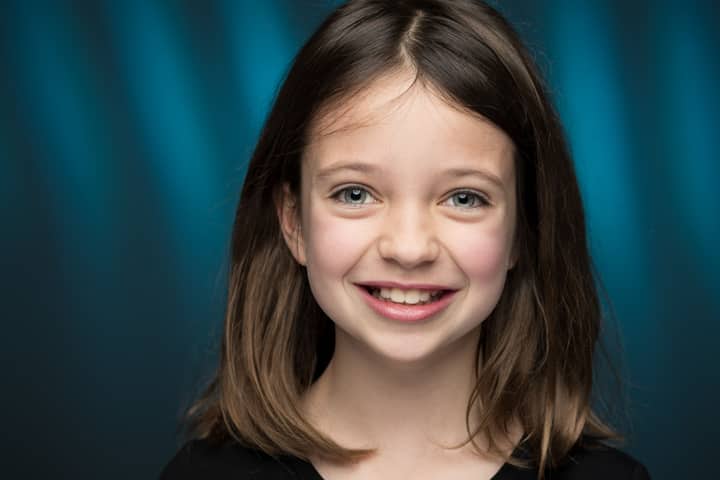 Portrait of charming little girl with light eyes and brown hair looking at camera against blue background