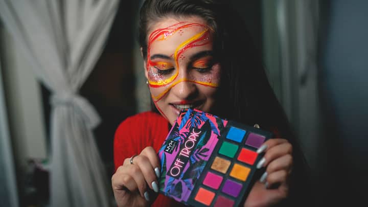 Young dreamy female with closed eyes and bright makeup demonstrating eyeshadow palette on blurred background indoors