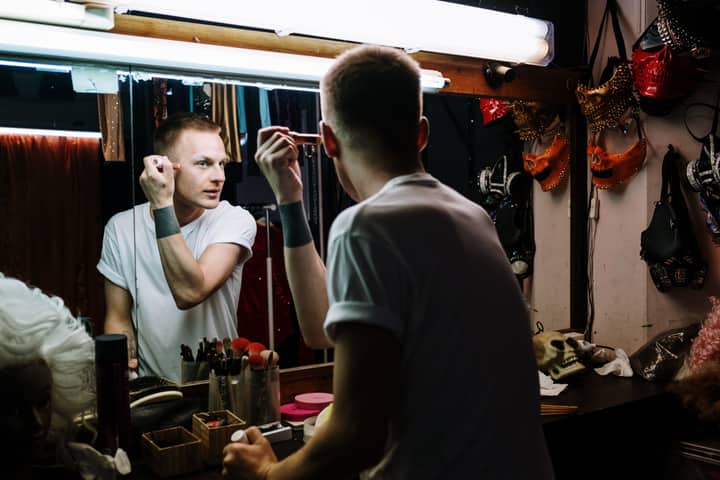 Drag performer applies makeup in a theater dressing room, reflecting transformation and identity.