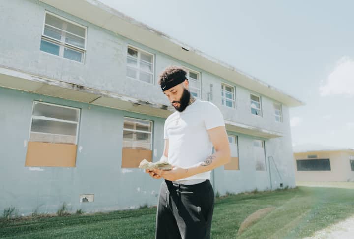A young man with facial hair counts money in front of a worn building on a sunny day.