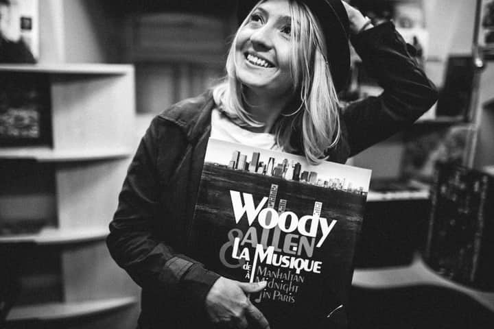 Black and white photo of a woman smiling and holding a vinyl record in a vintage shop.