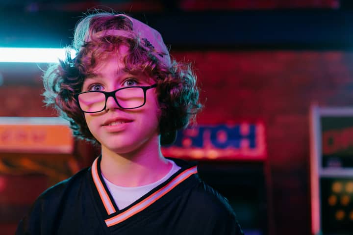 Teenager with glasses in neon-lit arcade, expressing joyful emotions and carefree spirit.