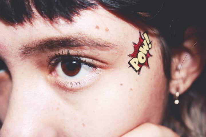 Detailed close-up of a brown eye with a colorful 'Pow' tattoo next to it.