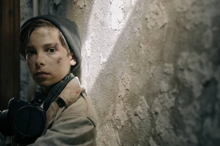 A young boy in a post-apocalyptic setting looking determined, wearing survival gear.