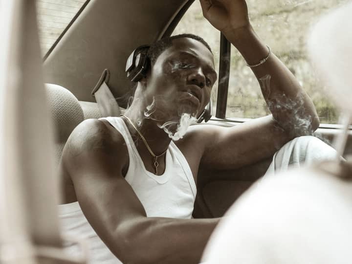 A young man in a car, wearing headphones and inhaling smoke, exudes a calm, introspective mood.