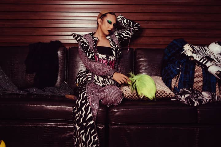 Young transgender male dancer in bright outfit and with makeup resting on leather couch tired after performance