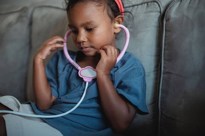 Side view of crop focused Asian kid wearing headband sitting on soft couch with toy stethoscope while playing doctor in cozy living room