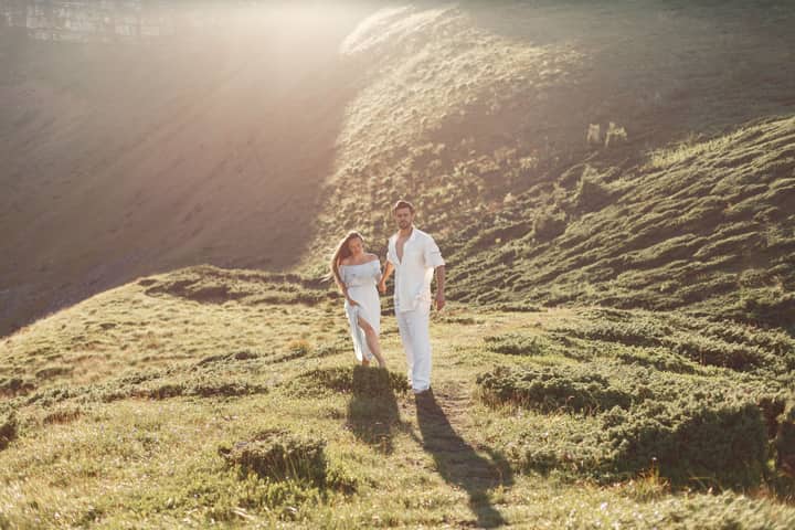 A couple in white clothes walking in a sunny grass field, enjoying a warm and serene moment.