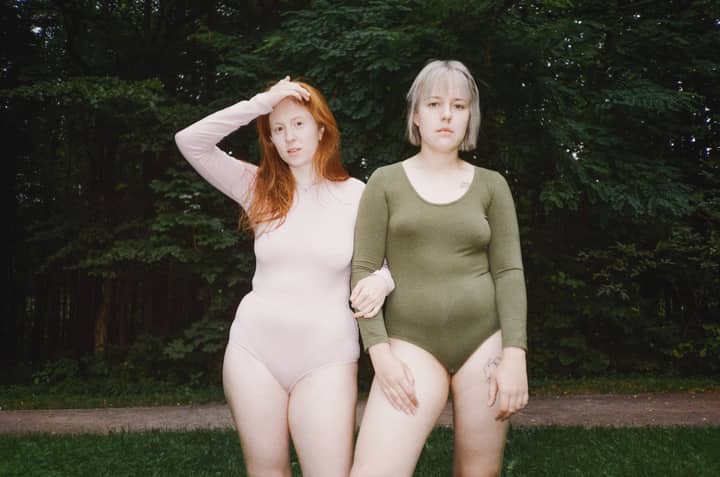 Two women in bodysuits embracing body positivity against a lush green backdrop.